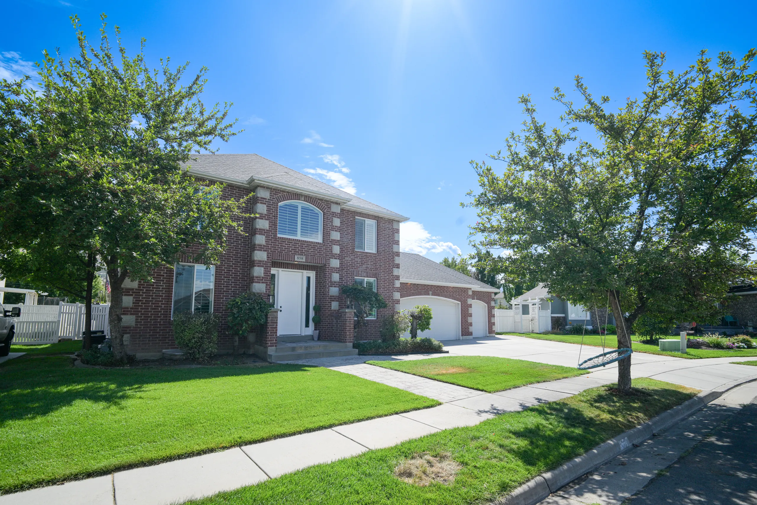 Two-story red brick home in Layton, Utah with a cut lawn, mature shade trees and a three-car garage, photographed from across the street